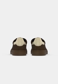 Brown suede sneakers with a beige textile heel accent, rounded toe, and rubber sole. Laced design with contrasting stitching details.