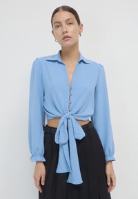 Blusa azzurro chiaro a maniche lunghe con colletto, bottoni frontali e dettaglio vita annodata. Tessuto morbido con una vestibilità rilassata e maniche arricciate.