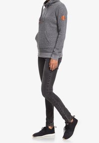 Felpa grigia con tasca frontale, cuciture tonali e patch in pelle sulla spalla. Abbinata a jeans neri slim e sneaker scure.