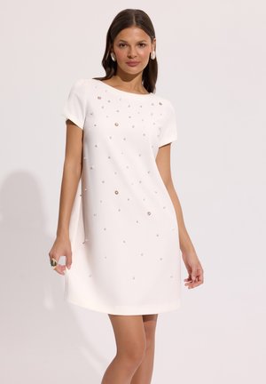 Kobieta w krótkiej białej sukience ozdobionej małymi perełkowymi koralikami, pozująca na tle jasnej, jednolitej ściany, z kolczykami i pierścionkiem.