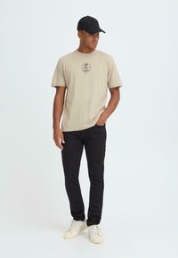 Bež pamučna majica s kružnim logom, uparena s crnim hlačama i svijetlim tenisicama. Model nosi crnu kapu.