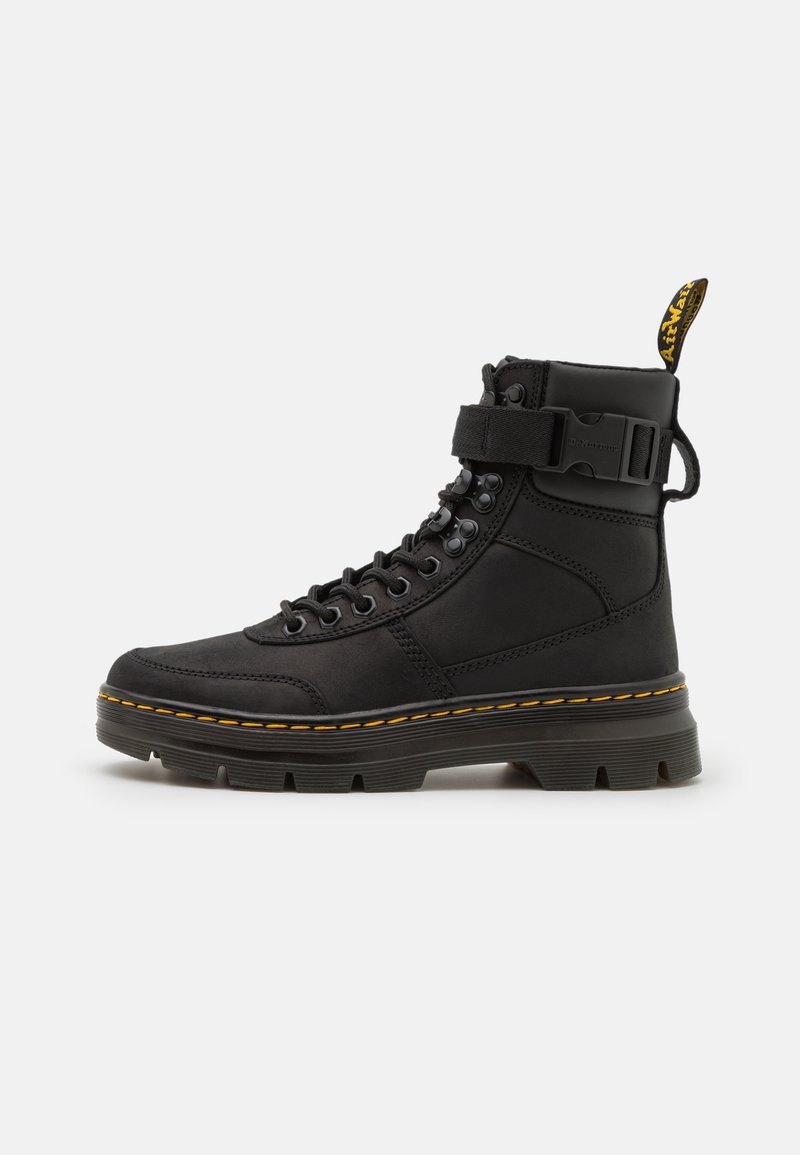 Bottine montante noire à lacets avec surpiqûres jaunes, sangle à boucle, semelle épaisse et languette de tirage portant la marque à l'arrière.