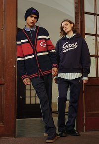 Cardigan bleu marine avec des rayures rouges et blanches et logo, associé à un pantalon de survêtement bleu marine. Le modèle porte un sweat à capuche bleu marine et une chemise à col à motifs.