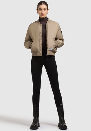 Khaki bomberjack met geribbelde manchetten en onderrand, voorrits en zijzakken, gedragen over een zwarte coltrui en legging met enkelboots.
