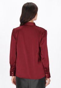 Bordowa jedwabna bluzka z wysokim kołnierzem, bufiastymi rękawami i zapinanymi mankietami; gładka tekstura i błyszczące wykończenie.