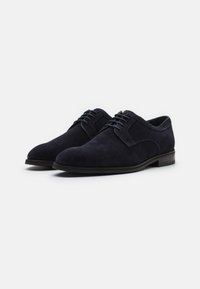 Scarpe di velluto blu scuro con punta arrotondata, lacci sul davanti e tacco basso. Presentano bordi lisci e una finitura testurizzata.