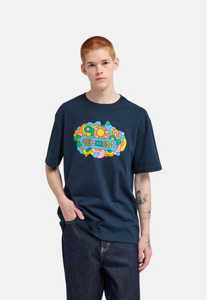 Młoda osoba z krótkimi czerwonymi włosami, ubrana w granatowy t-shirt z kolorowym abstrakcyjnym wzorem i tekstem "Element", w połączeniu z ciemnoniebieskimi dżinsami.