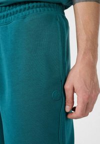 Teal joggers van zacht materiaal met een elastische tailleband, zijzakken en een klein reliëflogo op het linkerbeen.