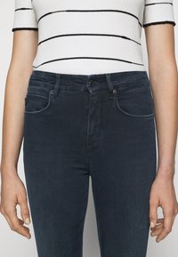 Jeans de mezclilla oscura con un diseño ajustado, bolsillos delanteros visibles y cierre de botón. La tela parece suave con un ligero estiramiento.