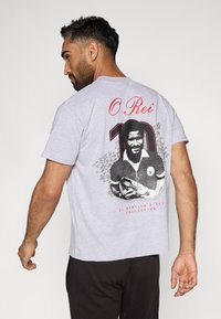 Γκρι βαμβακερό μπλουζάκι με στρογγυλή λαιμόκοψη. Στο πίσω μέρος υπάρχει μια ασπρόμαυρη εικόνα ενός άνδρα που κρατάει ένα παπούτσι ποδοσφαίρου, με τις λέξεις "Ο Ρέι" και "10" σε κόκκινο χρώμα.