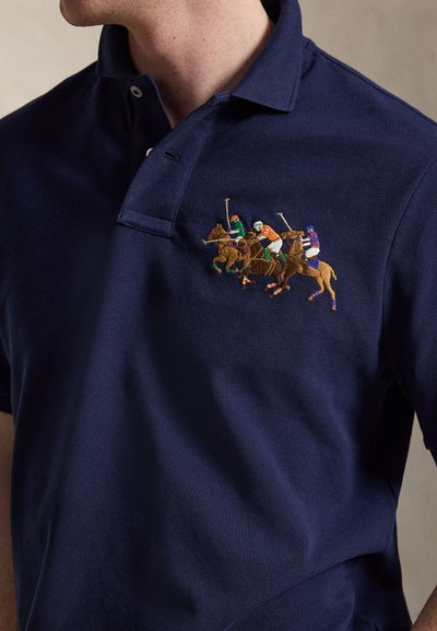 Navy plava polo majica s izvezenim logotipom tri igrača polo na konjima u akciji, svaki s obojenim kacigama i opremom.