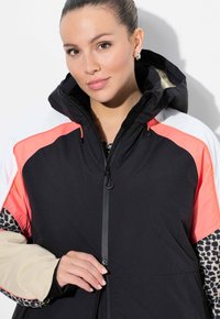 Černobílá bunda s kapucí s bloky v korálové a béžové barvě. Má zapínání na zip a vzory na vnitřní podšívce. Měkká textura.