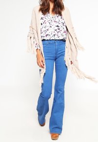 Cardigan beige en maille avec franges, porté sur un chemisier imprimé floral et un jean bleu évasé. Des bottines marron à talons complètent la tenue.