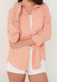 Chemise à boutons rayée avec des rayures verticales orange et blanches, manches longues retroussées, poche poitrine et coupe ample. Associée à un short blanc.