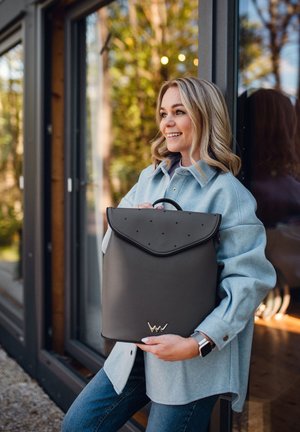 Grau lederner Rucksack mit einer runden Klappe, verziert mit durchbrochenen Akzenten und einem metallenen Logo. Hat eine strukturierte rechteckige Form.