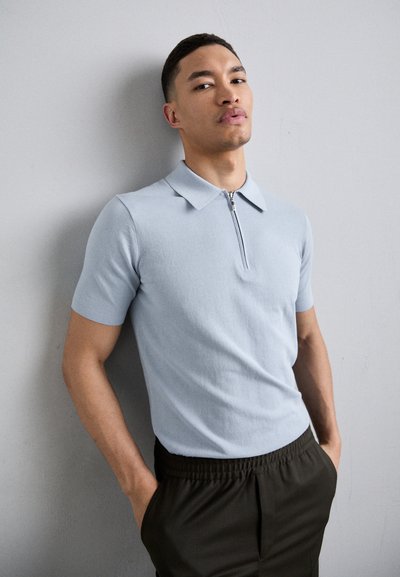 Polo shirt bleu pâle à manches courtes avec un col zippé, fabriqué en tissu lisse. Associé à un pantalon sombre à taille élastique. Vue à corps entier.