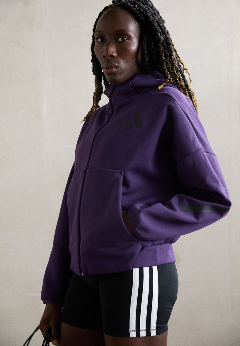 Lila zip-up hoodie i mjukt tyg med fickor och svarta accenter, i kombination med svarta shorts som har vita ränder på sidorna.