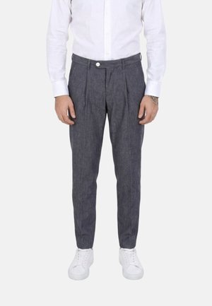 Uomo che indossa pantaloni plissettati grigio scuro, camicia bianca con bottoni e sneaker bianche, in piedi contro uno sfondo chiaro uniforme.