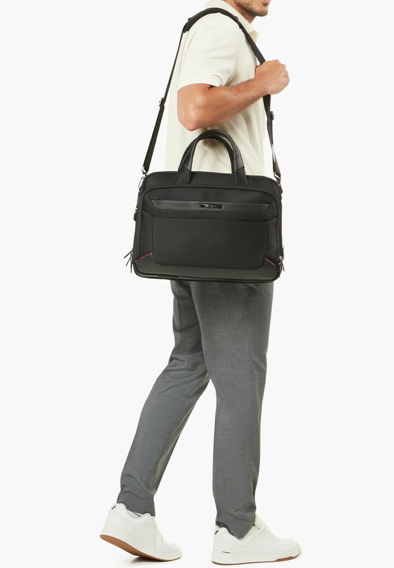 Man carrying a black laptop bag with handles and shoulder strap, wearing white sneakers, gray pants, and a light short-sleeve shirt.