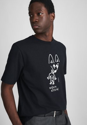 Junger Mann in einem schwarzen Maison Kitsuné T-Shirt mit einem weißen abstrakten Fuchs- und Menschenfigur-Graphic, eingesteckt in graue Hosen mit einem schwarzen Gürtel.