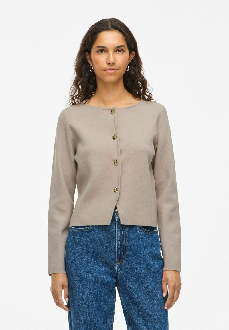 Femme aux cheveux sombres ondulés portant un cardigan beige à boutons et un jean taille haute bleu, se tenant devant un fond blanc uni.