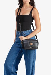 Borsa a tracolla in pelle nera con chiusura in metallo dorato, superficie testurizzata e logo in rilievo. Indossata con pantaloni in denim a vita alta e una maglietta nera.