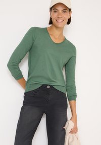 Grüner langärmeliger Pullover mit Rundhalsausschnitt und gerippten Bündchen, kombiniert mit schwarzen Jeans. Der Pullover hat einen abgerundeten Saum und eine weiche Textur.