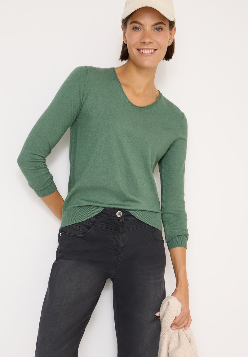 Grüner langärmeliger Pullover mit Rundhalsausschnitt und gerippten Bündchen, kombiniert mit schwarzen Jeans. Der Pullover hat einen abgerundeten Saum und eine weiche Textur.