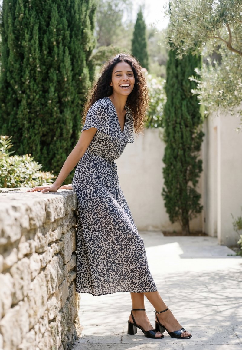 Femme aux cheveux bouclés sourit en étant assise sur un mur en pierre, portant une robe imprimé léopard et des sandales noires à talons dans un jardin ensoleillé.