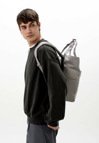 Grå ryggsäck med flera fack, rektangulär form och justerbara remmar. Bärs över en mörk sweatshirt med en slät yta.