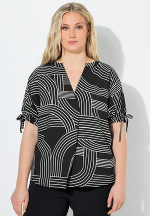 Lyshåret kvinde iført en sort og hvid bluse med geometrisk mønster og korte ærmer bundet ved armene samt sorte bukser, stående mod en hvid væg.