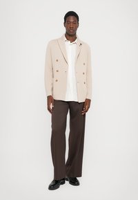 Beige Doppelreiher-Blazer mit braunen Knöpfen über einem weiß gestreiften Hemd, kombiniert mit braunen weiten Hosen und schwarzen Schuhen.