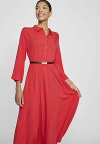 Raudona suknelė ilgomis rankovėmis su apykakle, turinti mygtukų užsegimą ir įliemenuotą juosmenį su juodu diržu. Lygus audinys, tekanti siluetas.