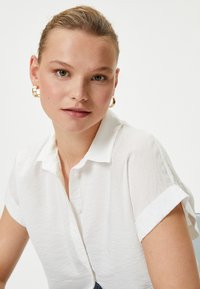 Witte button-up shirt met korte opgerolde mouwen, gemaakt van een textuurstof. Het model draagt gouden hoepeloorbellen en heeft een neutrale make-uplook.