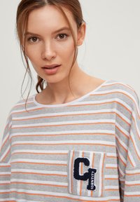 Maglia a maniche lunghe a righe grigie e bianche con dettagli arancioni, dotata di una toppa in feltro blu navy con "C" e "IDENTITY" sulla tasca del petto.