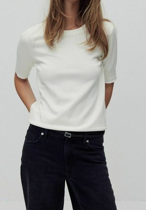 Kobieta ubrana w biały krótki top włożony do wysokich czarnych jeansów z czarnym paskiem, stojąca z rękami za plecami.