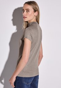 Beige short-sleeve top with a high neckline and subtle shimmer, paired with blue jeans. Texture is smooth with a fitted design.