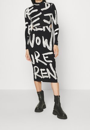 Kobieta w czarnej, midi sukience z odważnym, białym nadrukiem i wycięciami w talii, w połączeniu z czarnymi, masywnymi botkami za kostkę.