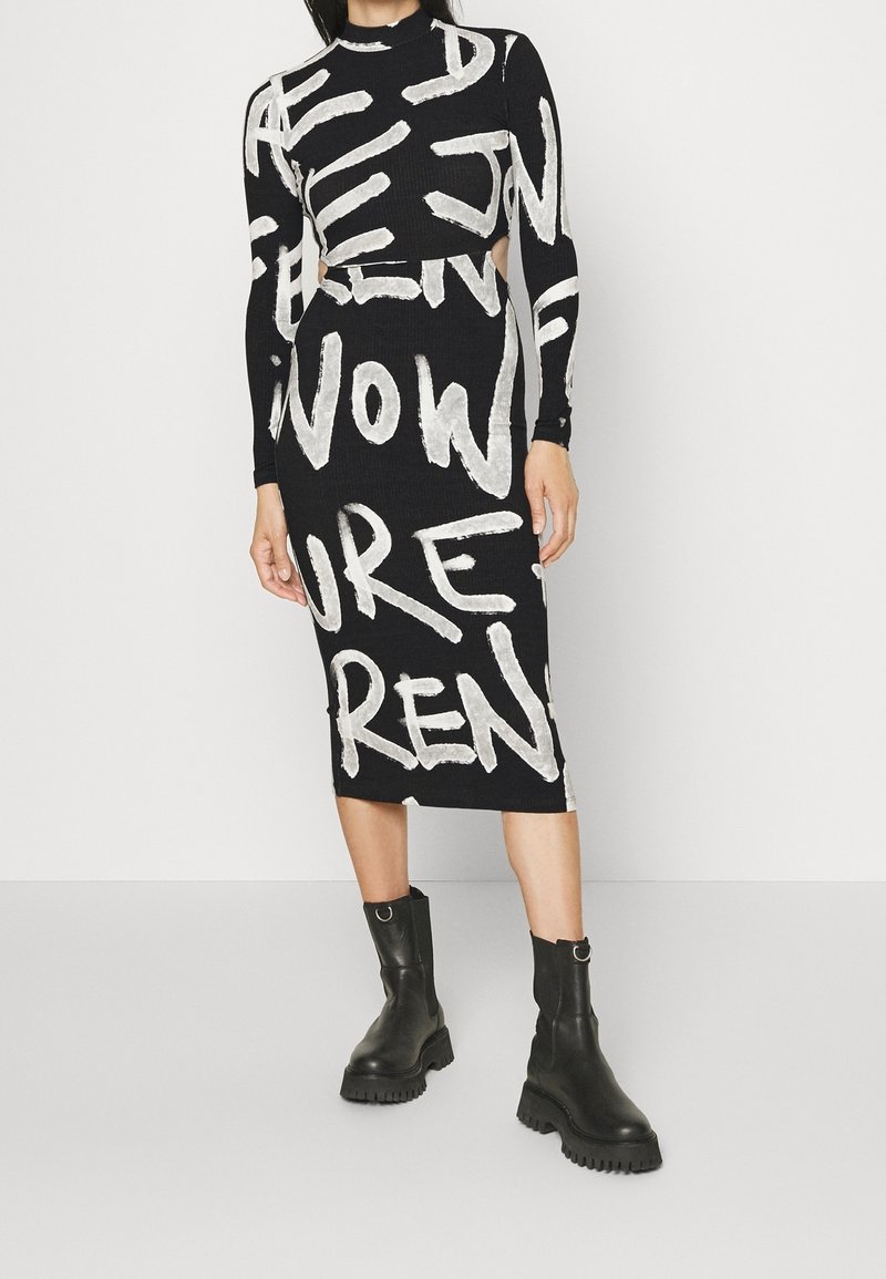 Femme portant une robe midi noire avec un texte graphique blanc audacieux et des découpes à la taille, associée à des bottines noires à semelle épaisse.