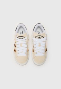 Beige sneakers med hvite snører, med svart merkevare og en leopardmønstret aksent. Mykt semsket skinnmateriale og rund tådesign.