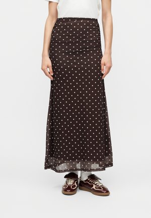 Kobieta ubrana w długą ciemnobrązową spódnicę w białe kropki, białe skarpetki, ciemnobrązowe trampki i biały krótki top z krótkim rękawem.