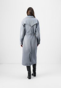 Manteau long en denim bleu clair avec une taille ceinturée, un col pointu, un empiècement triangulaire et des fentes latérales, associé à des bottes noires jusqu'au genou.
