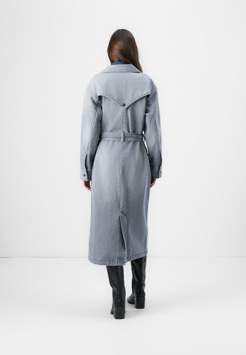 Himmelblauer Denim-Langmantel mit tailliertem Gürtel, spitzem Kragen, dreieckigem Schulterbereich und seitlichen Schlitzen, kombiniert mit schwarzen kniehohen Stiefeln.