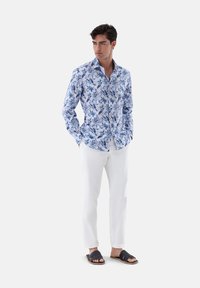 Camicia a maniche lunghe con fantasia floreale blu, abbinata a pantaloni bianchi e sandali neri, in piedi su uno sfondo neutro.