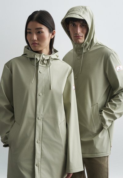 Deux mannequins portant des imperméables à capuche vert olive clair, se tenant devant un fond gris clair uni, les mains dans les poches.