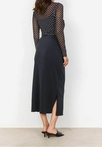 Sort, knælang nederdel med en sideslids, parret med en gennemsigtig, langærmet top med hvide prikker, båret med lavhælede sorte mules.