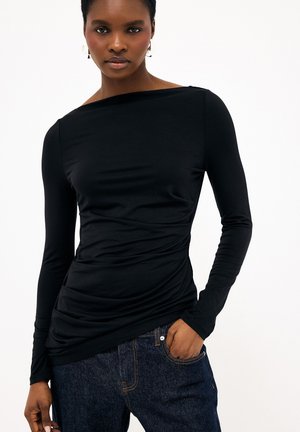 Ženska, oblečena v oprijet zgornji del s črnimi dolgimi rokavi z rahlim nabiranjem, združen z temno modrimi jeansi, pozira z eno roko v žepu.