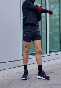 Athlète en tenue de sport noire et gants rouges étire son bras en plein air près d’un bâtiment moderne en verre.
