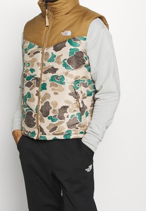 Mann trägt eine The North Face Camouflage-Steppweste mit beigefarbenem Schulterbereich über einem hellgrauen Sweatshirt und schwarzen Hosen mit Logo am linken Oberschenkel.