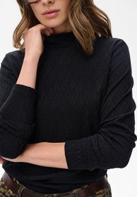 Kobieta w czarnej, fakturowanej bluzce z długim rękawem i wysokim kołnierzem, trzymająca kołnierz jedną ręką, z brązowym paskiem, w widocznych spodniach w cętki ze wzorem lamparta.